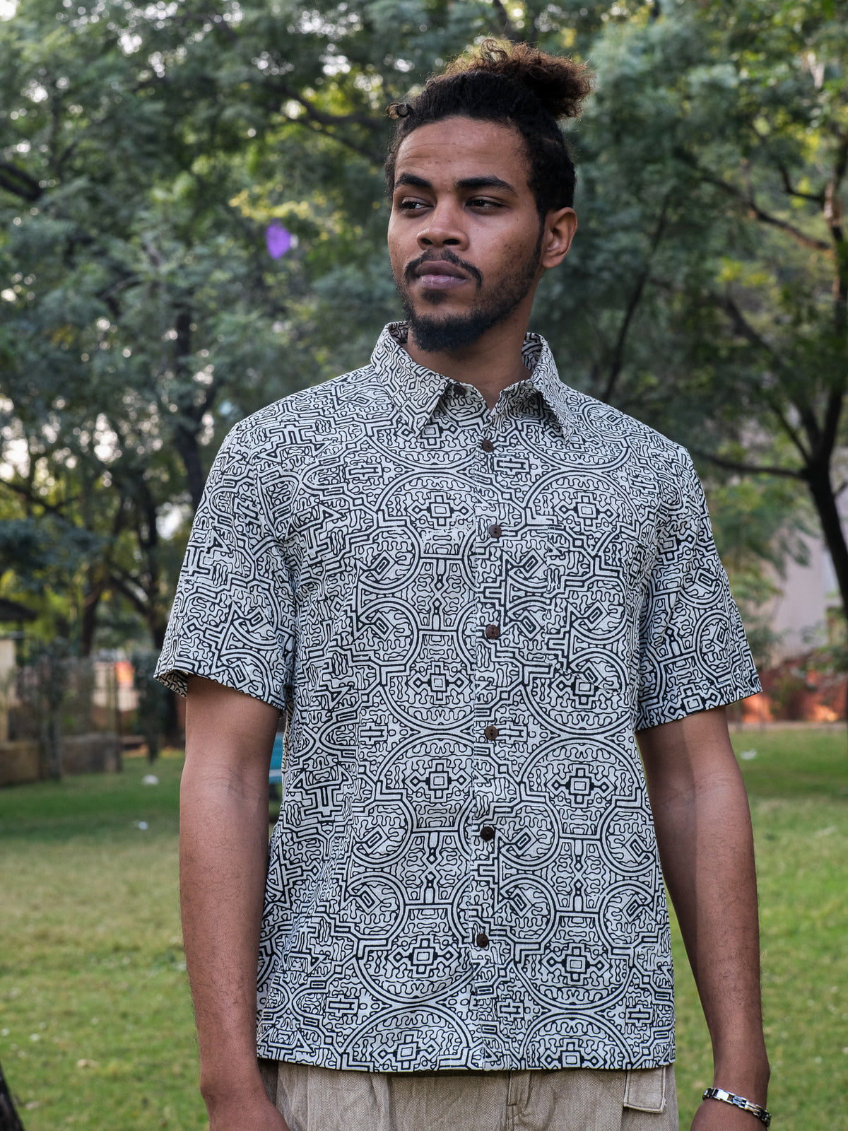 A man with a beard and tied-back hair stands outdoors, surrounded by trees and grass, wearing the white Shipibo Shirt—a short-sleeved cotton shirt adorned with an intricate Shipibo print.