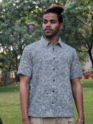 A man with a beard and tied-back hair stands outdoors, surrounded by trees and grass, wearing the white Shipibo Shirt—a short-sleeved cotton shirt adorned with an intricate Shipibo print.