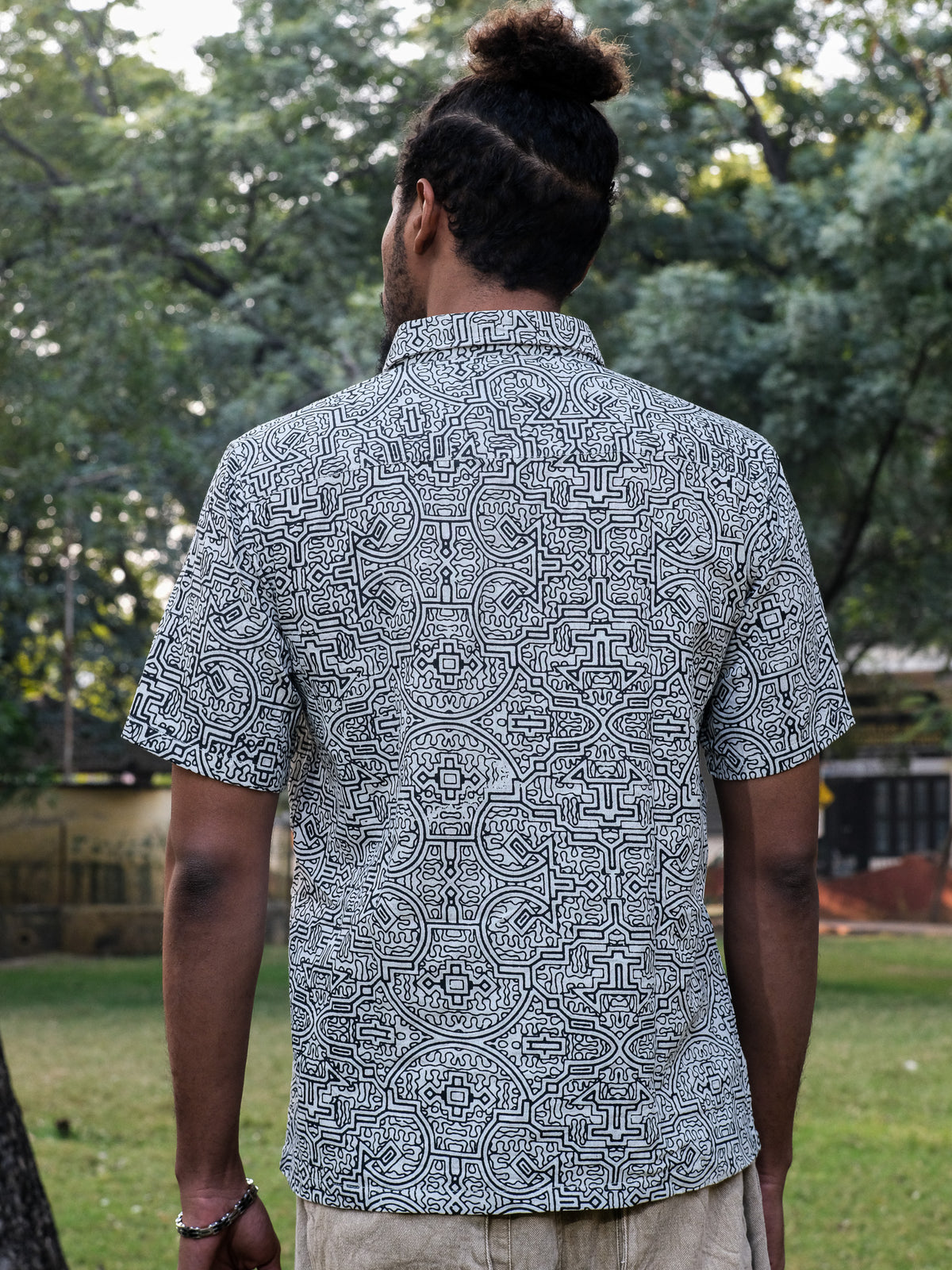 A person with a bun hairstyle, seen from behind, wears a short-sleeved white Shipibo Shirt featuring a distinctive Shipibo print while standing outdoors in a grassy area with trees in the background.