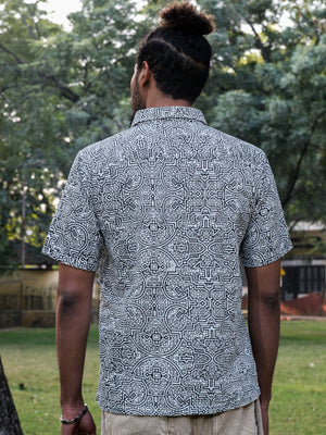 A person with a bun hairstyle, seen from behind, wears a short-sleeved white Shipibo Shirt featuring a distinctive Shipibo print while standing outdoors in a grassy area with trees in the background.