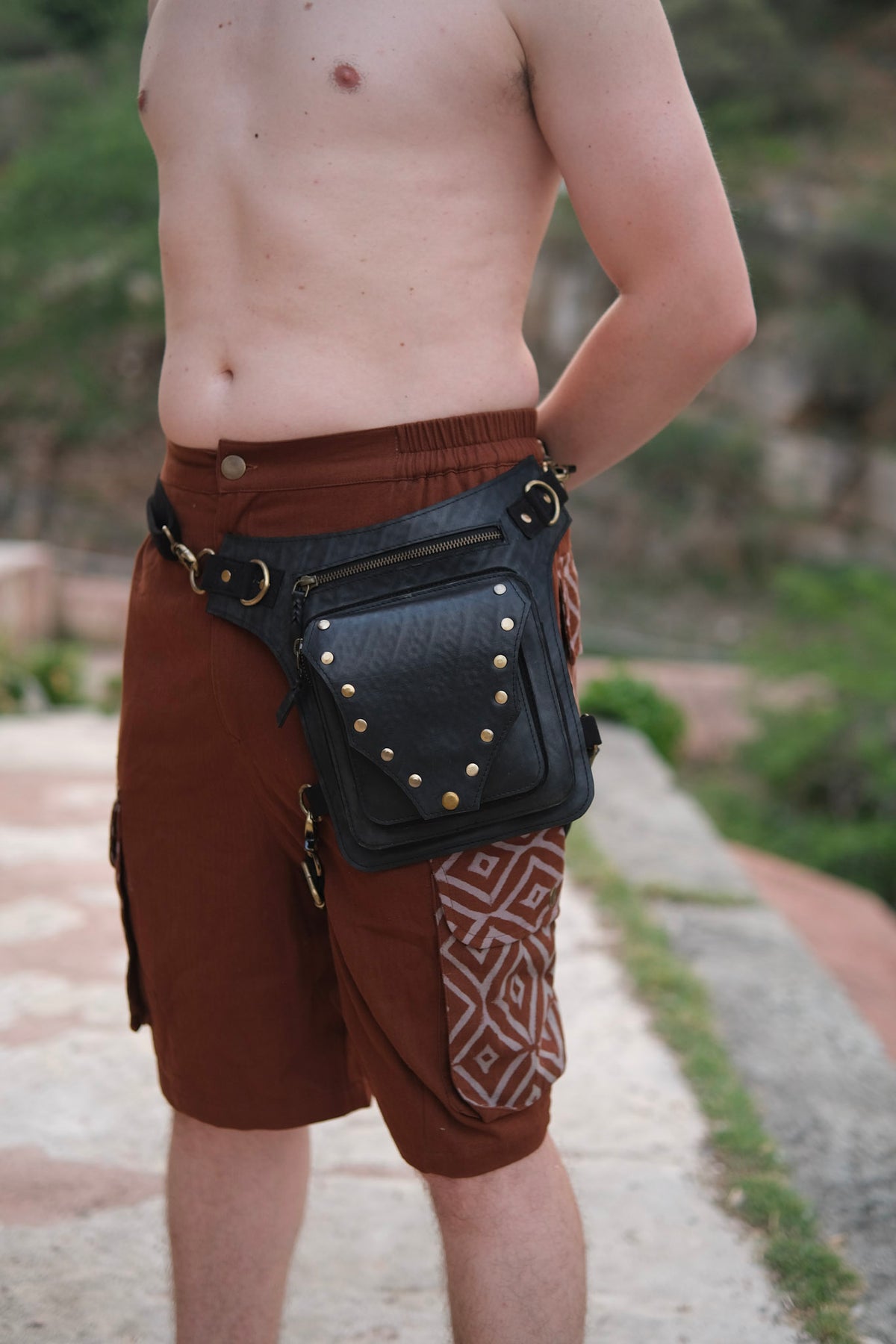 A black, eco-friendly Nomad Fest Reclaim Bag featuring brass studs and zippered compartments, worn at the hip over brown shorts as a sustainable festival accessory.