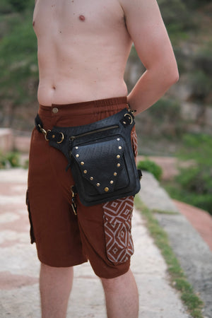 A black, eco-friendly Nomad Fest Reclaim Bag featuring brass studs and zippered compartments, worn at the hip over brown shorts as a sustainable festival accessory.