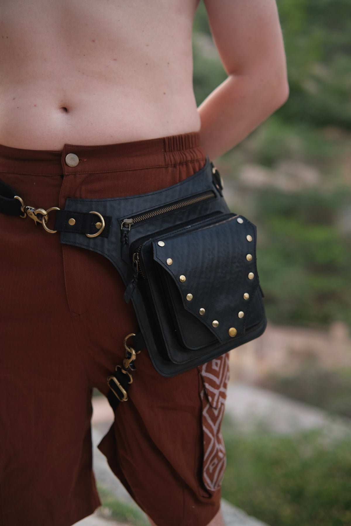 Nomad Fest Reclaim Bag: a black hip bag with zippered pockets and brass studs, worn around the waist with a clip attachment to the thigh as a sustainable festival accessory.