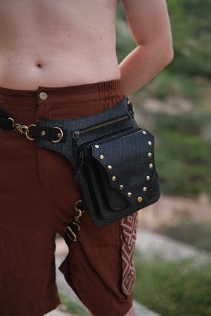Nomad Fest Reclaim Bag: a black hip bag with zippered pockets and brass studs, worn around the waist with a clip attachment to the thigh as a sustainable festival accessory.