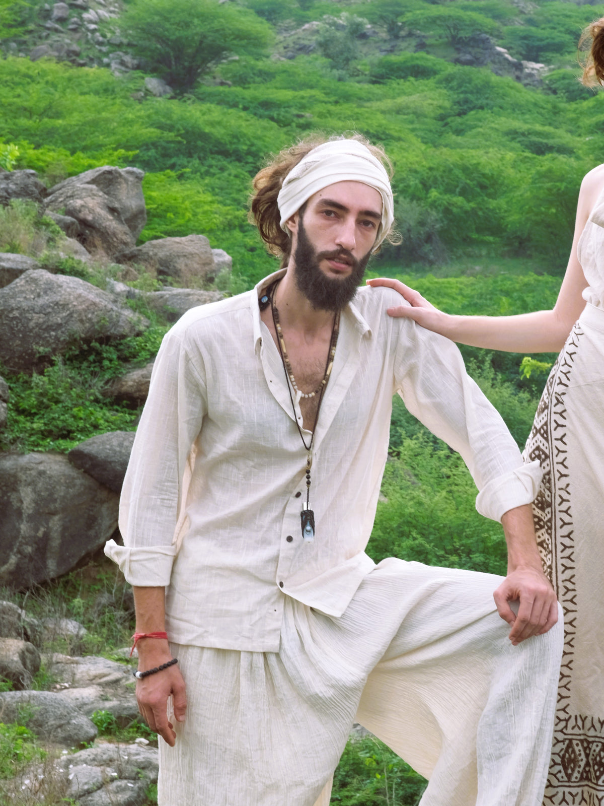A bearded man in a white YinYang Shirt and headscarf poses outdoors among greenery and rocks, while another person’s hand rests on his shoulder.