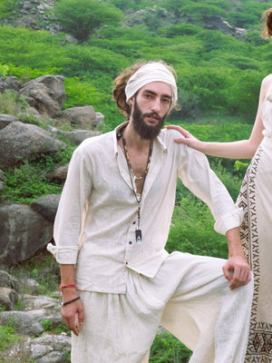 A bearded man in a white YinYang Shirt and headscarf poses outdoors among greenery and rocks, while another person’s hand rests on his shoulder.