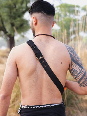 A shirtless individual stands outdoors with the Luca Festival Bag on their shoulder. They have a short haircut, a beaded necklace, and a tattoo on their left arm. Blurred trees and tall grass in the background highlight the festival vibe.