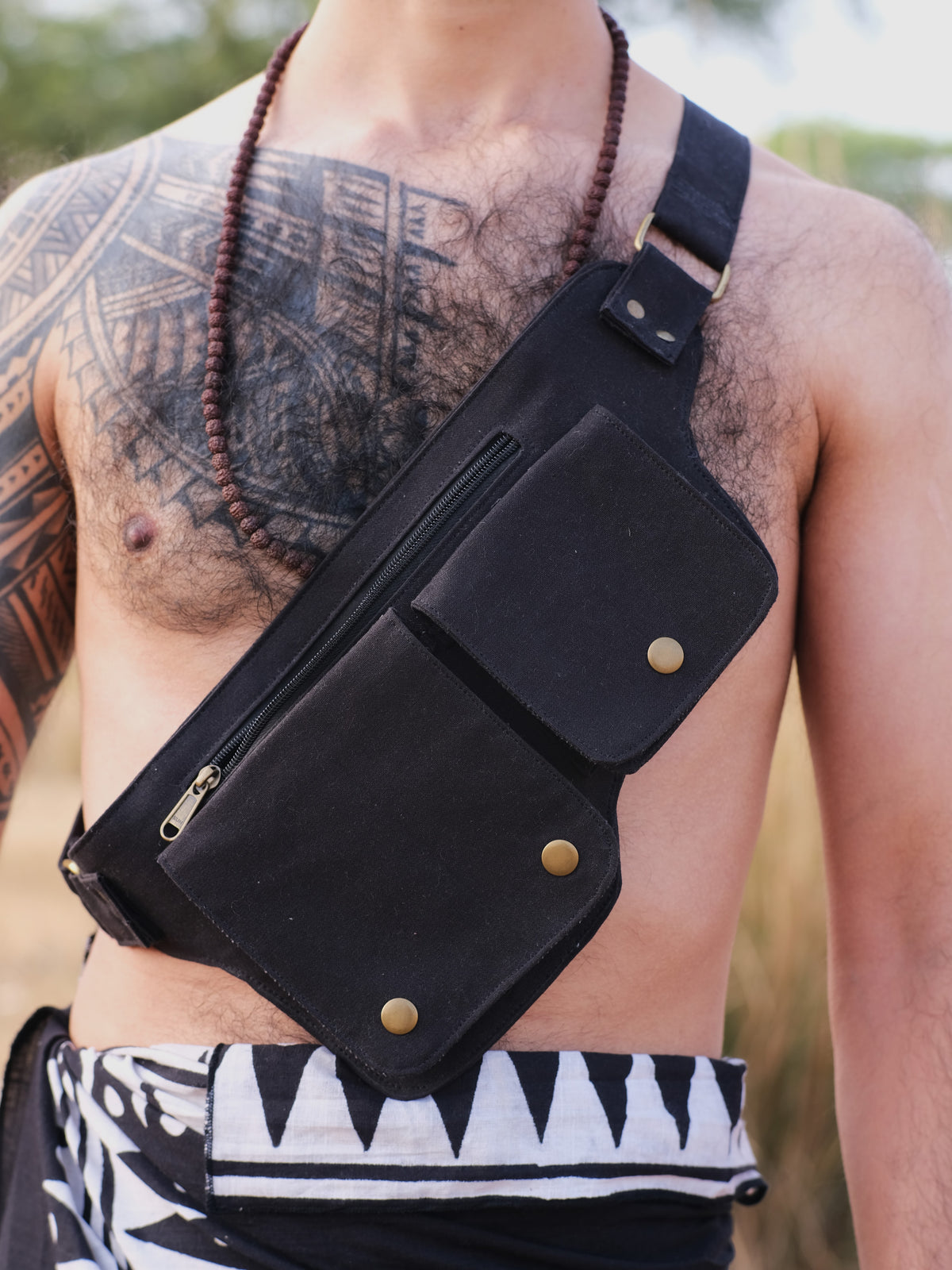 A shirtless person highlights the premium black Luca Festival Bag with dual metal-snapped pockets, complementing their chest tattoo and beaded necklace, while wearing patterned black and white pants against a blurry green backdrop, capturing a festival vibe.