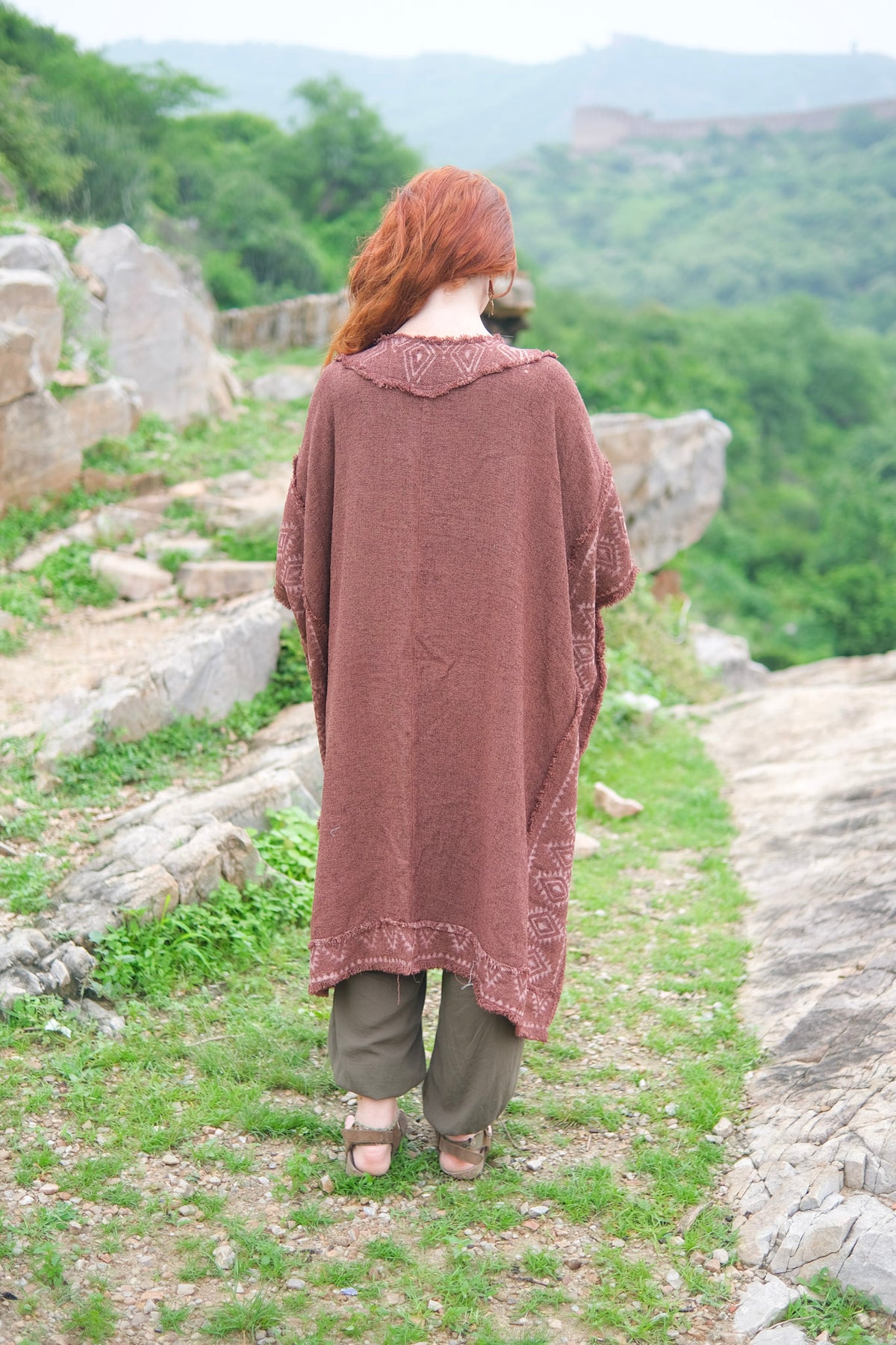 Back view of the model walking away in the brick red knit kimono, showing the plain knit back, the block printed collar and the cobweb trim along the hem, over olive harem pants on a stone path with green hills behind.