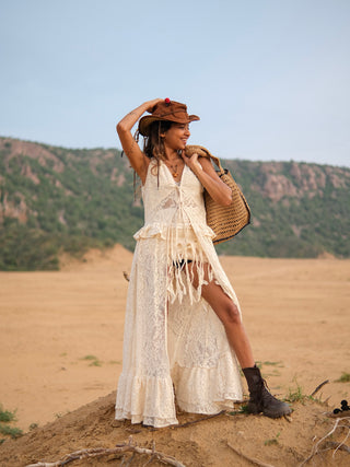 Wearing the Niraya Desert Shrug over an ivory lace dress and boots, a woman stands on sandy terrain, holding a hat and a woven bag to effortlessly channel festival style against a backdrop of hills and clear sky.