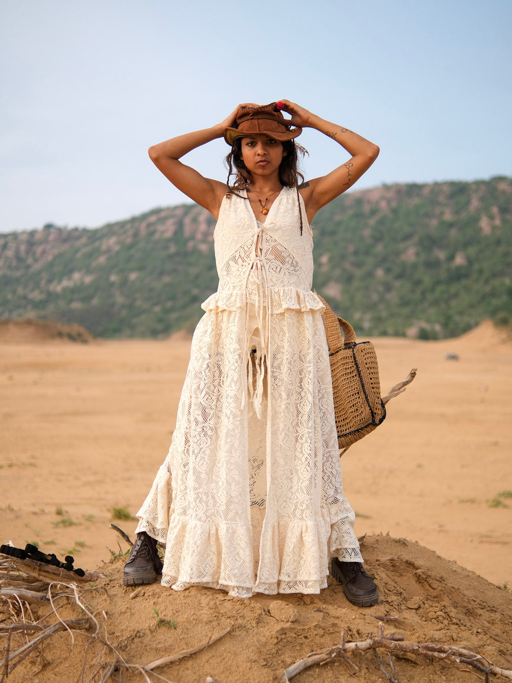 Wearing the Niraya Desert Shrug in natural, a woman stands on sandy ground, holding a brown hat on her head. Embracing effortless festival styling, she pairs the shrug with a long cream lace dress, carries a woven bag, and wears boots, with hills and greenery in the background.