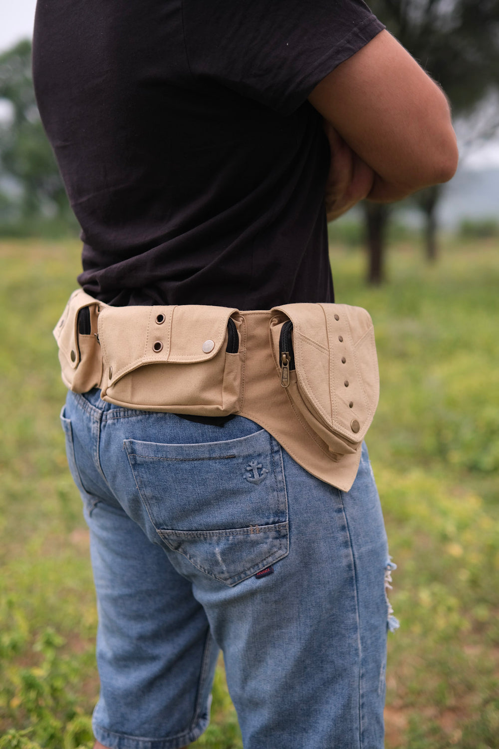 A person wearing a black shirt and denim shorts stands outdoors with arms crossed, showing the Verdant Multi-Pocket Utility Belt Bag—featuring several handy pouches—worn around their waist. Slung casually over one shoulder is a cotton canvas bag with tribal geometric accents and an adjustable strap.