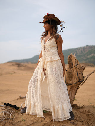 A woman in a cream lace dress and a brown feathered hat stands in a desert landscape beside a woven bag and driftwood, the Niraya Desert Shrug draped elegantly around her shoulders as she gazes into the distance, embodying effortless festival style.