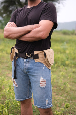 A person stands outdoors with arms crossed, wearing a black t-shirt, ripped denim shorts, and the Verdant Multi-Pocket Utility Belt Bag adorned with tribal geometric accents. The background is grassy with trees and hills.