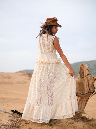 A woman in a long, sleeveless ivory lace dress and brown hat stands in a sandy desert, holding her skirt. Channeling effortless festival style, she completes her look with the Niraya Desert Shrug. A woven bag hangs on a weathered tree stump beside her, with sparse hills visible in the background.