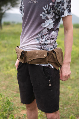 A person wearing a floral t-shirt, black shorts, and the Verdant Multi-Pocket Utility Belt Bag—crafted from brown cotton canvas with an adjustable strap and tribal geometric accents—stands outdoors on grass with a blurred natural background.