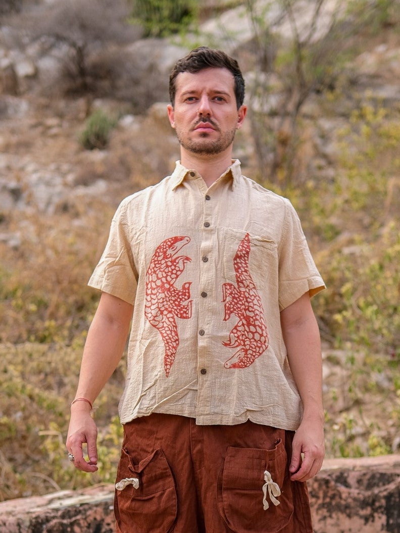 A person stands outdoors wearing the beige Pangolin shirt, featuring two red animal designs, paired with brown pants against a rocky, dry background with sparse vegetation.