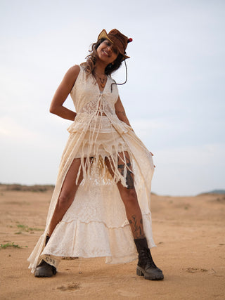 Wearing the Niraya Desert Shrug draped effortlessly around their shoulders, a person in a white bohemian-style dress with delicate ivory lace accents and a brown cowboy hat stands smiling on sandy terrain. Showcasing festival styling with chunky boots, they reveal visible tattoos on their legs beneath an overcast sky.