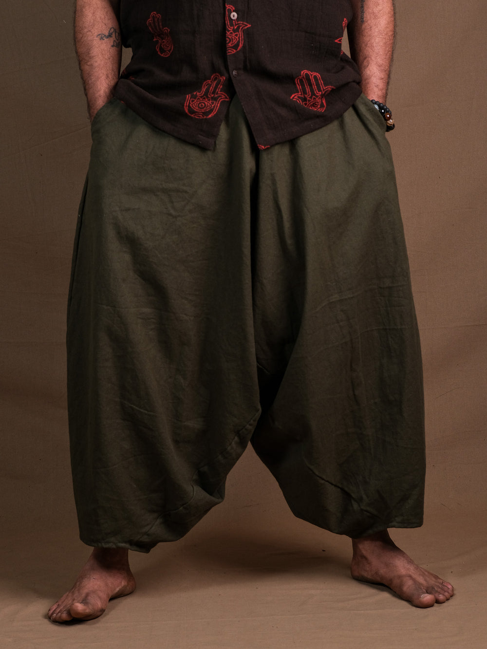A person stands barefoot in green Sirwal pants, hands in pockets, wearing a dark shirt with red handprint designs. The loose-fitting pants and bold top add a modern twist to their look against a plain brown background.