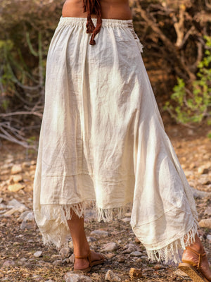 Back view of the model in the natural cream Nirvighna Skirt on a rocky path, showing the gathered elasticated waistband, the plain cotton back panel and the raw-fringed layered asymmetrical hem.
