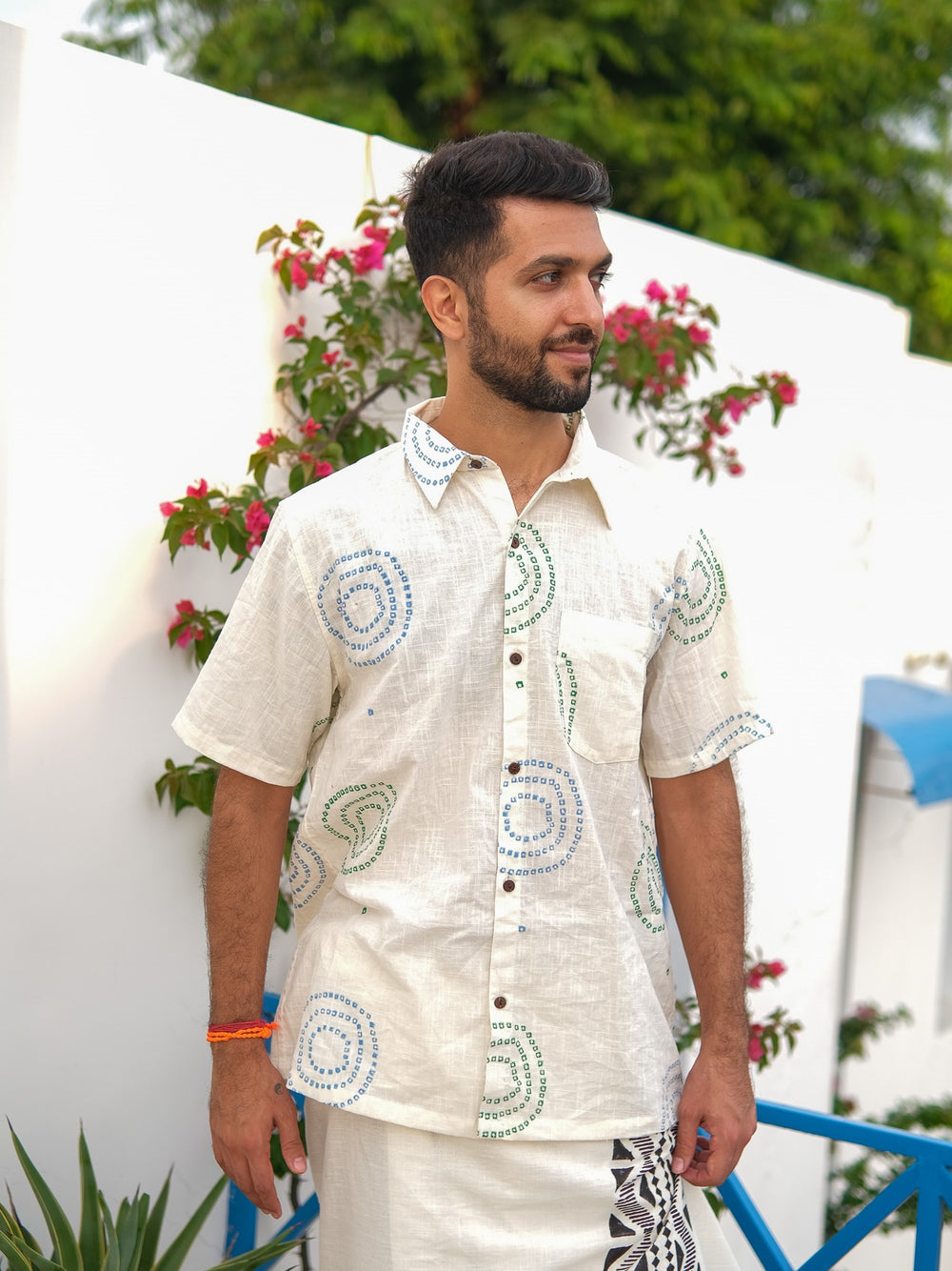 A man with a beard stands outdoors, wearing the Ishya Shirt—a light-colored, patterned button-up made from premium fabric—and a white skirt. Pink flowers and green leaves grow on a white wall behind him, with a blue railing visible to the right.