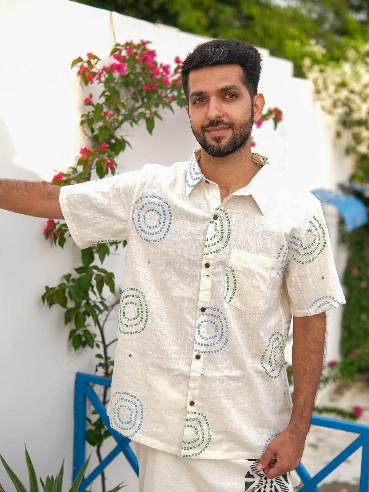 A man with short dark hair and a beard stands outdoors, dressed in the Ishya Shirt—a premium white short-sleeve shirt featuring blue and green circular patterns. Behind him, pink flowers and greenery decorate a white wall.