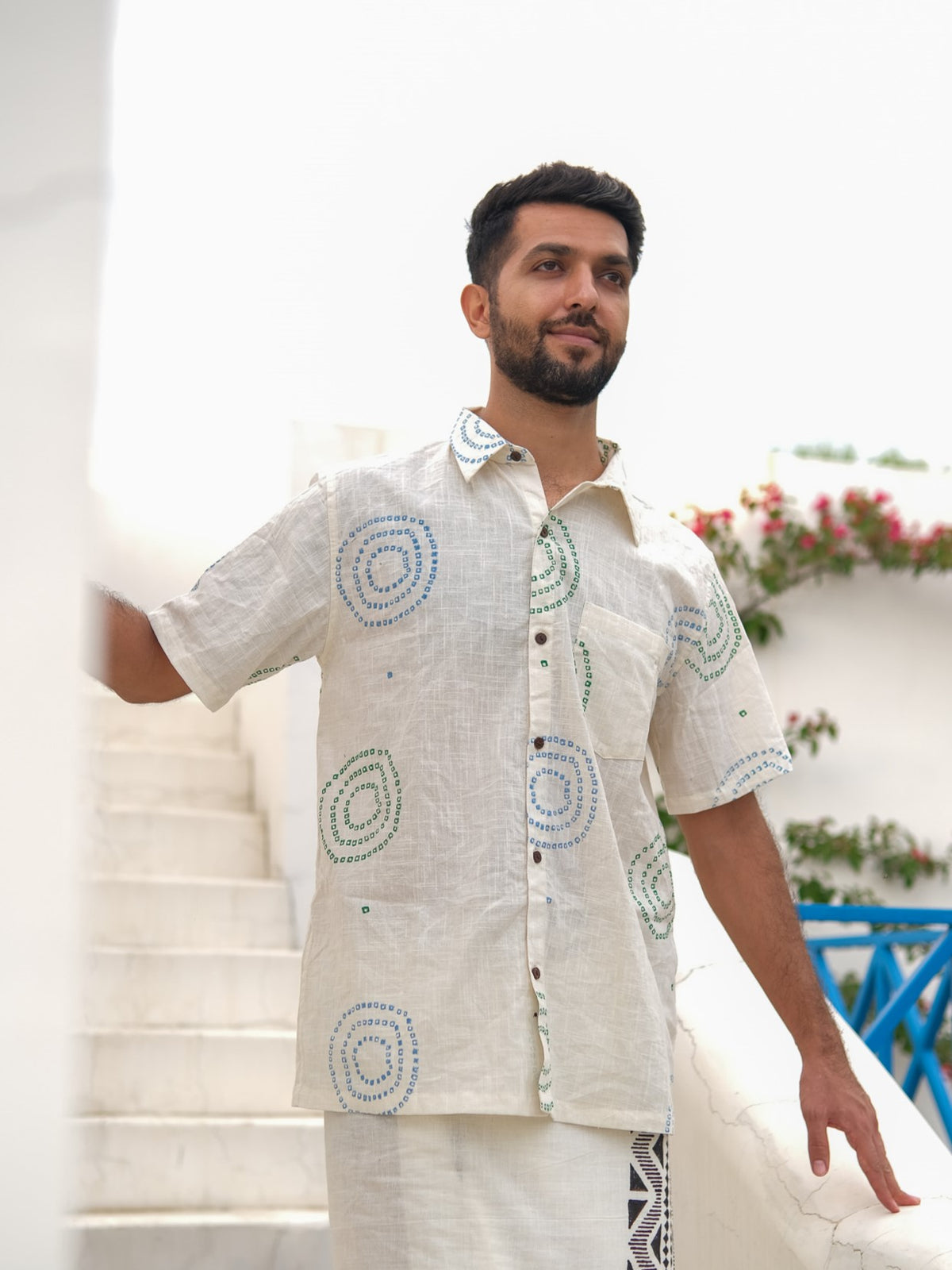 A man with a short beard stands on white stairs, wearing the Ishya Shirt—a short-sleeved shirt featuring a light base adorned with circular block print patterns—as flowers and a blue railing provide a charming backdrop.