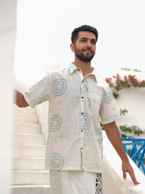 A man with a short beard stands on white stairs, wearing the Ishya Shirt—a short-sleeved shirt featuring a light base adorned with circular block print patterns—as flowers and a blue railing provide a charming backdrop.