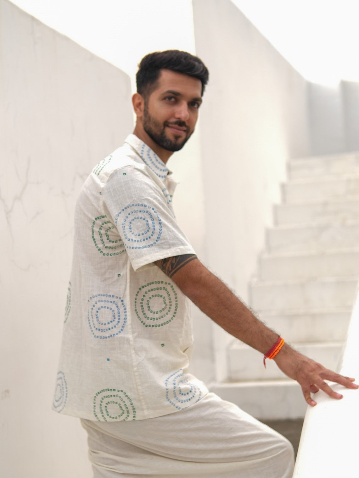 A man wearing the Ishya Shirt, a short-sleeve shirt featuring block print patterns and crafted from premium fabric, stands sideways on white stairs, gazing at the camera with a slight smile while resting his hand on a railing.