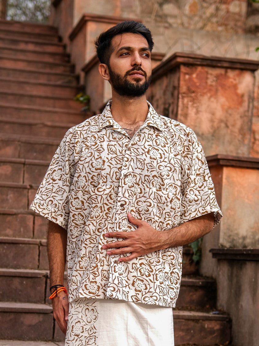 A bearded man stands on outdoor stone steps, dressed in the Aramya Baggy Shirt—a patterned, short-sleeved shirt crafted from premium quality fabric—and light-colored pants. He gazes to the side with one hand resting on his stomach and the other hanging relaxed by his side.