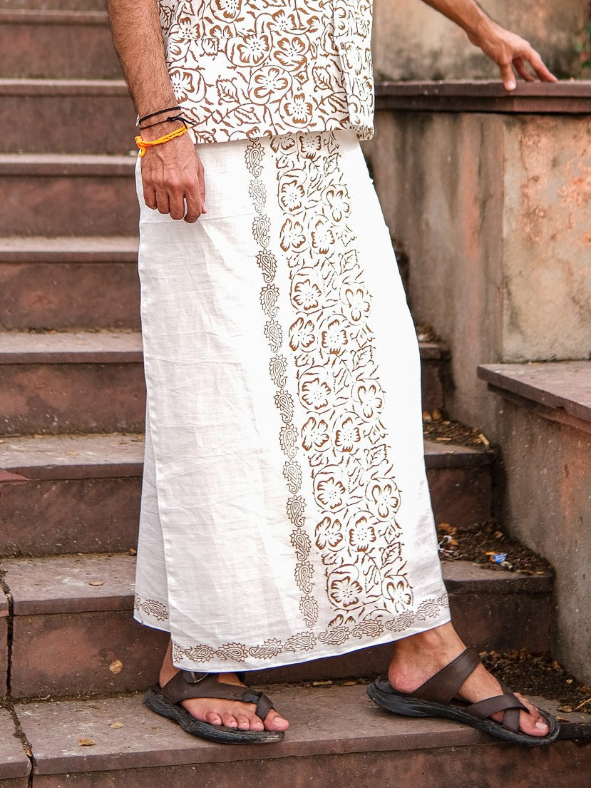 A person wearing the Vetra Dhoti, featuring a white block print with a floral pattern, and sandals stands on stone steps with one hand resting on a wall. Only the lower half of the body is visible.