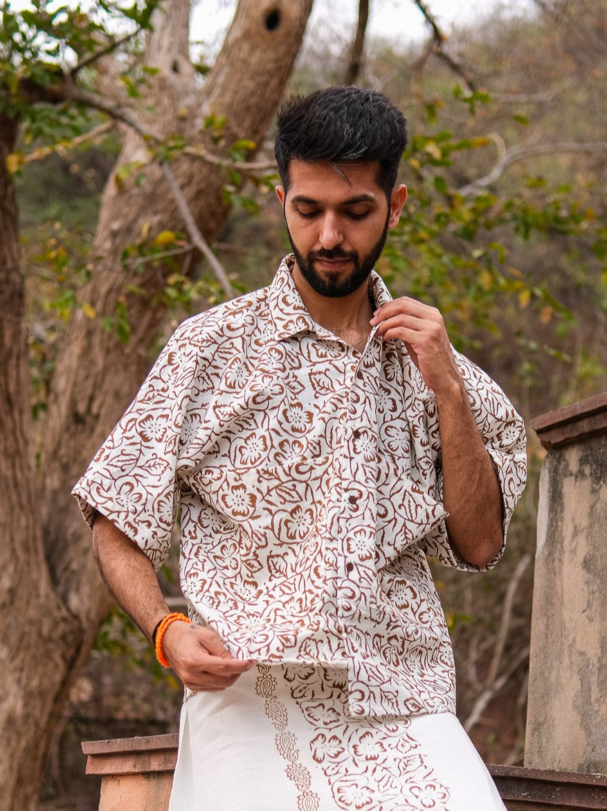 A man stands outdoors, looking down and holding the collar of his Aramya Baggy Shirt, which features a distinctive pattern. He pairs the shirt with white clothing, while trees and a stone structure form the backdrop.