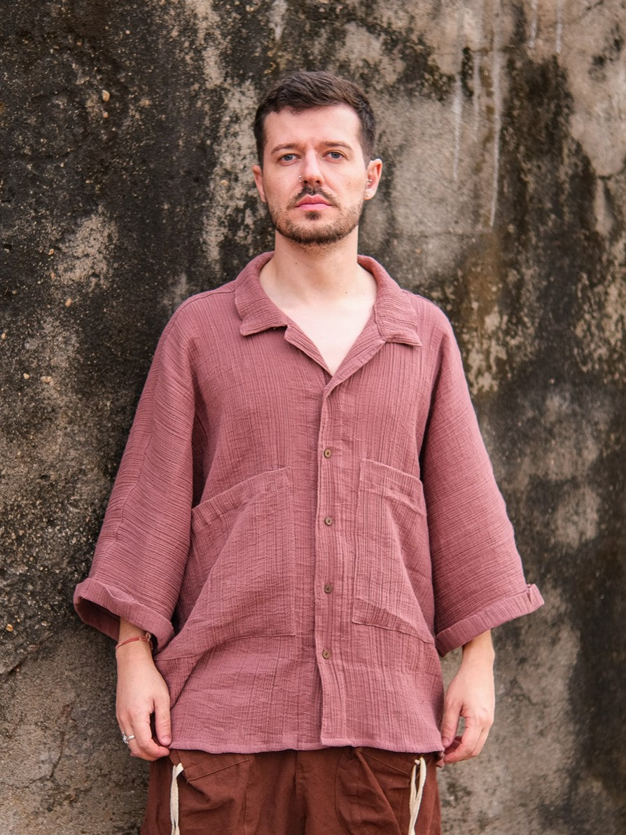 A man with short hair and a beard stands before a weathered wall, wearing the Yugma Baggy Shirt paired with brown pants for a relaxed, stylish look.