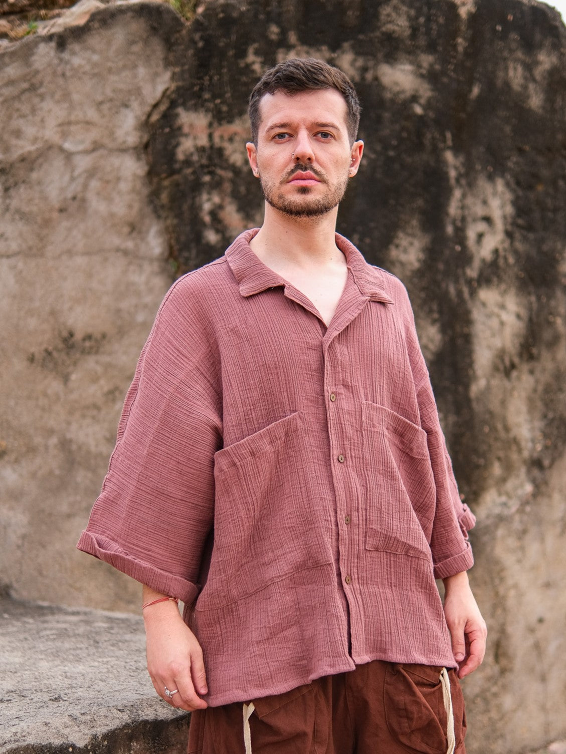A man wearing the Yugma Baggy Shirt with large pockets and brown pants stands outdoors by a stone wall, looking at the camera.