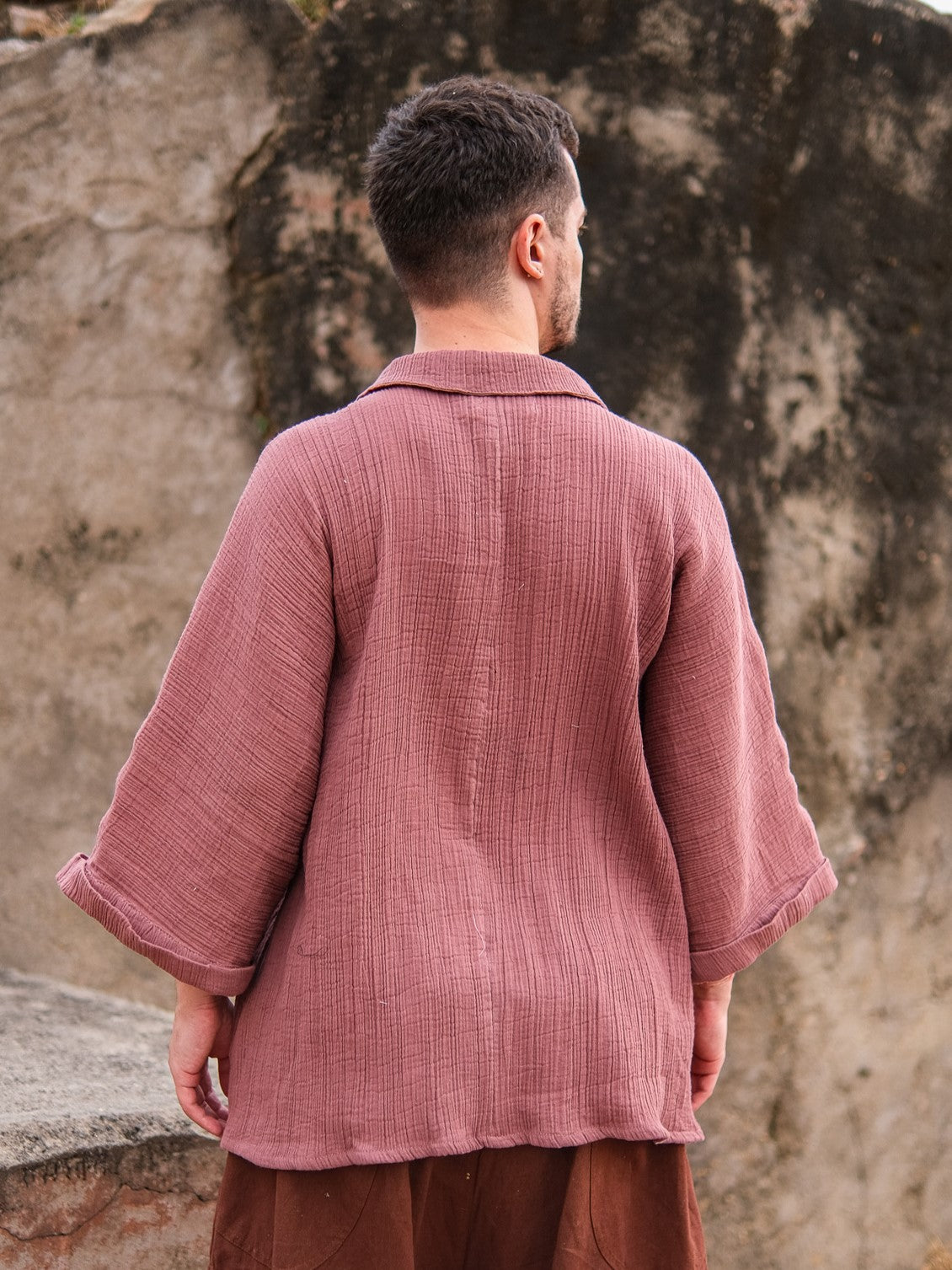 A person stands outdoors with their back to the camera, wearing a loose Yugma Baggy Shirt made from premium quality fabric and brown pants, set against a textured stone or concrete wall.