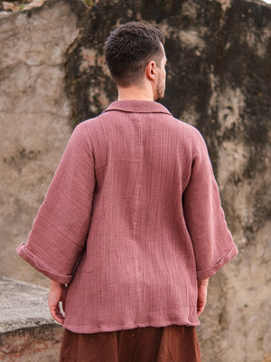 A person stands outdoors with their back to the camera, wearing a loose Yugma Baggy Shirt made from premium quality fabric and brown pants, set against a textured stone or concrete wall.