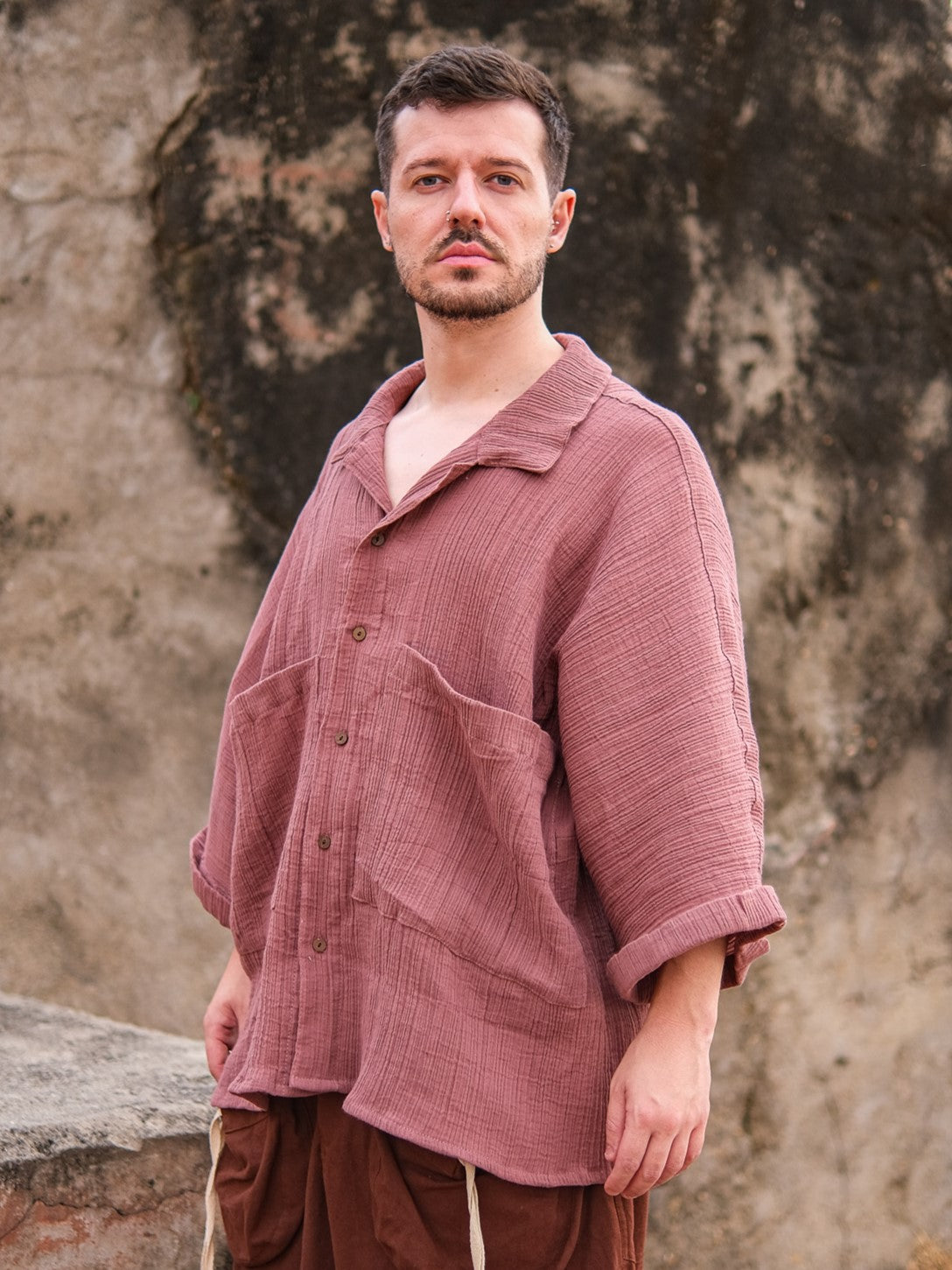 A man with short brown hair and light facial hair stands outdoors by a stone wall, wearing the Yugma Baggy Shirt—crafted from premium quality fabric with large pockets, rolled-up sleeves, and an elegant design.
