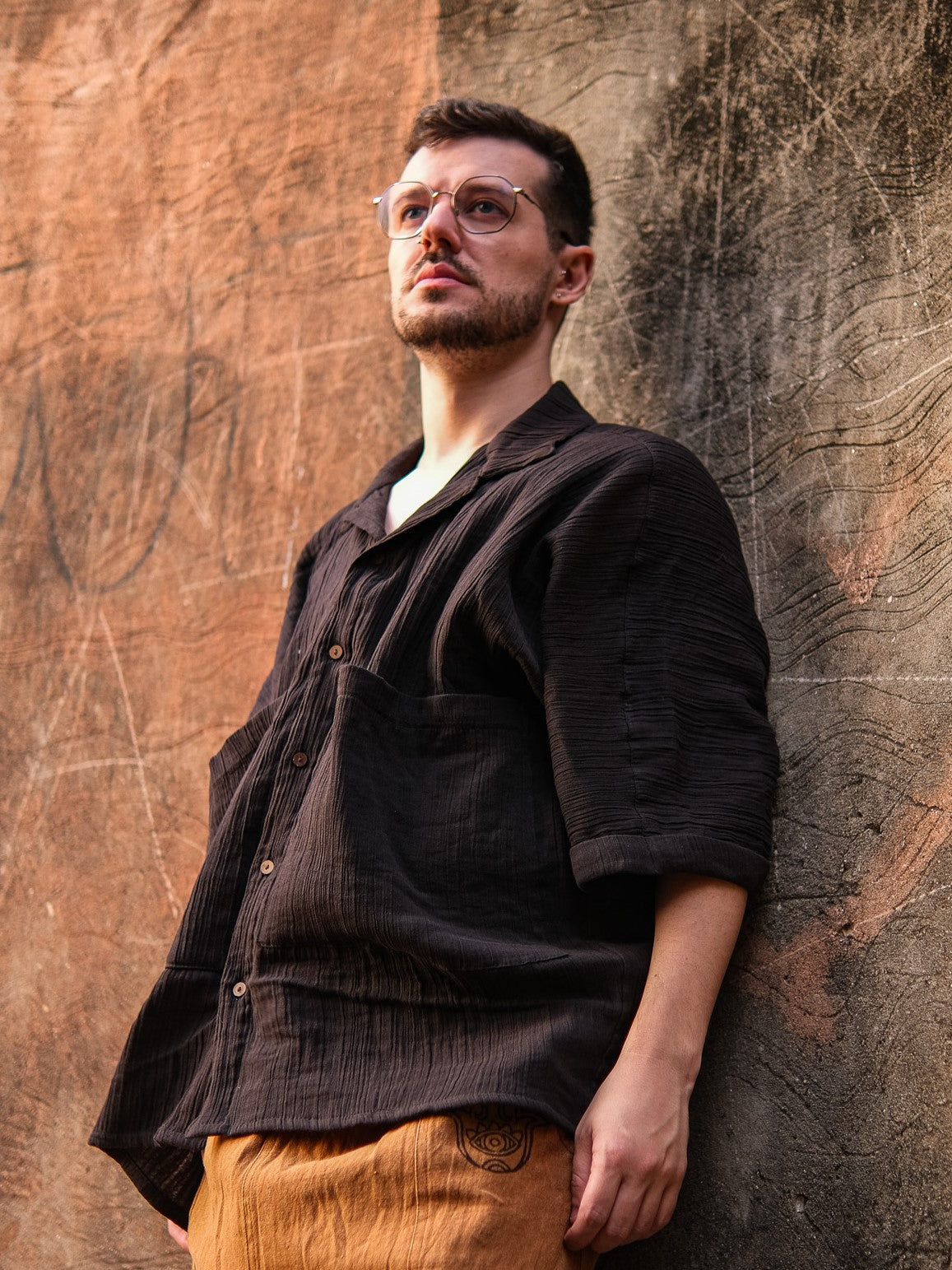 A man with short hair, glasses, and a beard stands by a textured wall wearing the Yugma Baggy Shirt and tan pants, gazing slightly upward with a thoughtful look.