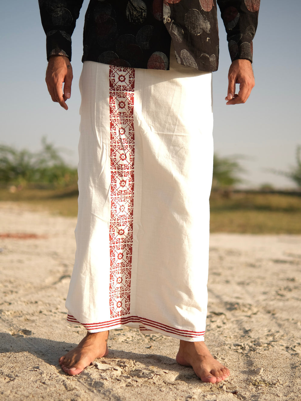 A person standing barefoot on sand, wearing the stylish Chakrot Dhoti made from high-quality fabric, which features a white garment with red patterns down the front and is paired with a black shirt adorned with circular designs. Greenery and sky are visible in the background.