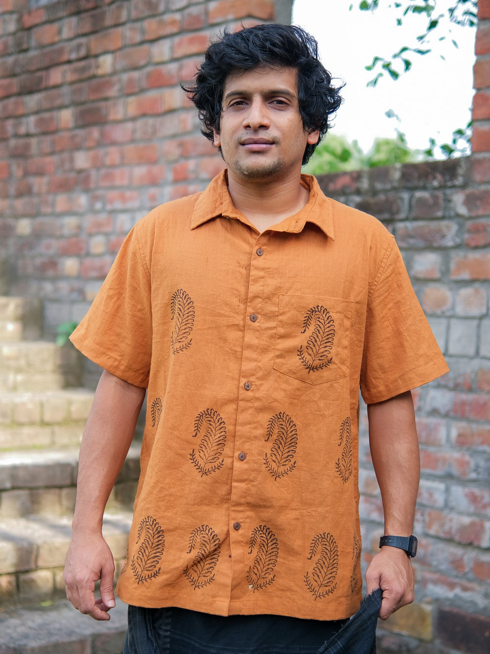 A person with wavy dark hair wears the Udaya Shirt—an elegant men’s orange short-sleeved shirt with a subtle leaf pattern—standing outdoors in front of brick walls and stone steps.
