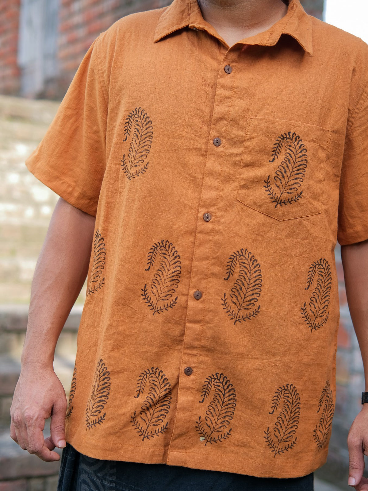 A man wearing the Udaya Shirt, a short-sleeved, light brown button-up with an elegant black leaf pattern, stands outdoors near brick steps.