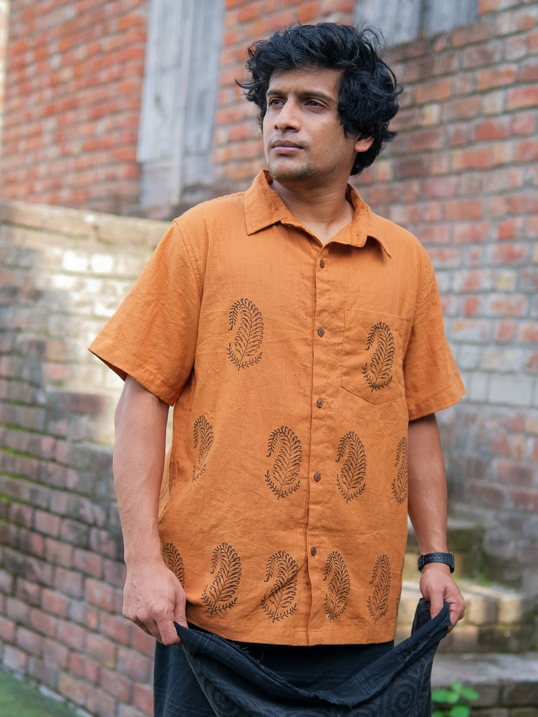A man stands outdoors beside a brick wall, wearing the Udaya Shirt—an orange, short-sleeved men’s shirt adorned with a paisley pattern. He gazes to the side with a neutral expression and holds a black cloth in his hands.