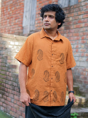 A man stands outdoors beside a brick wall, wearing the Udaya Shirt—an orange, short-sleeved men’s shirt adorned with a paisley pattern. He gazes to the side with a neutral expression and holds a black cloth in his hands.