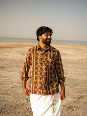 A man stands on a sandy beach wearing the Varun Handblock Printed Cotton Shirt—a testament to Indian craftsmanship—and a white traditional garment, smiling and looking to the side. The sea and sky are visible in the background.
