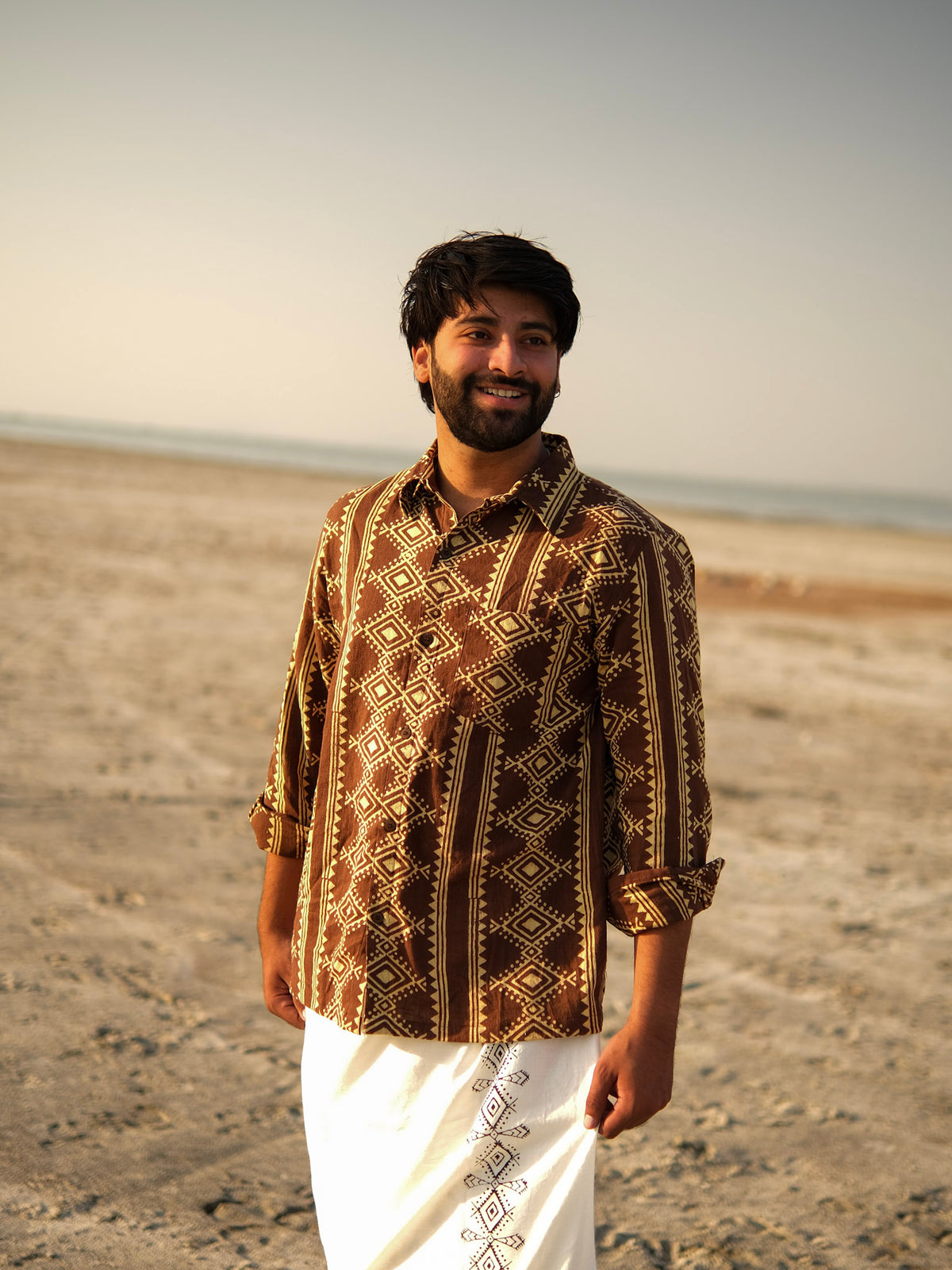Wearing the Varun Handblock Printed Cotton Shirt in a patterned brown design paired with a white traditional garment, a man stands smiling on a sandy beach with the ocean and sky behind him—a beautiful showcase of Indian craftsmanship.