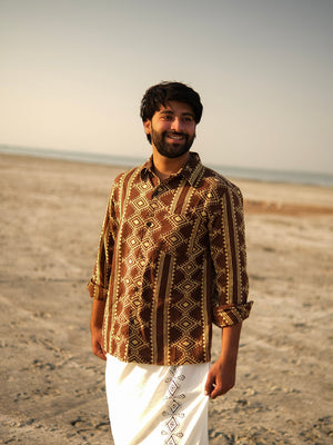 Wearing the Varun Handblock Printed Cotton Shirt in a patterned brown design paired with a white traditional garment, a man stands smiling on a sandy beach with the ocean and sky behind him—a beautiful showcase of Indian craftsmanship.