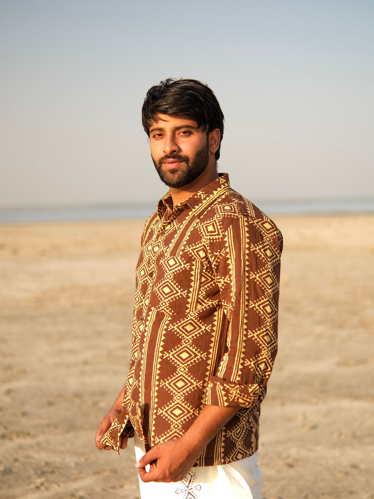 A man with dark hair and a beard stands on a sandy beach, wearing the Varun Handblock Printed Cotton Shirt that reflects exquisite Indian craftsmanship. The sky is clear and the background reveals a peaceful, empty shoreline.