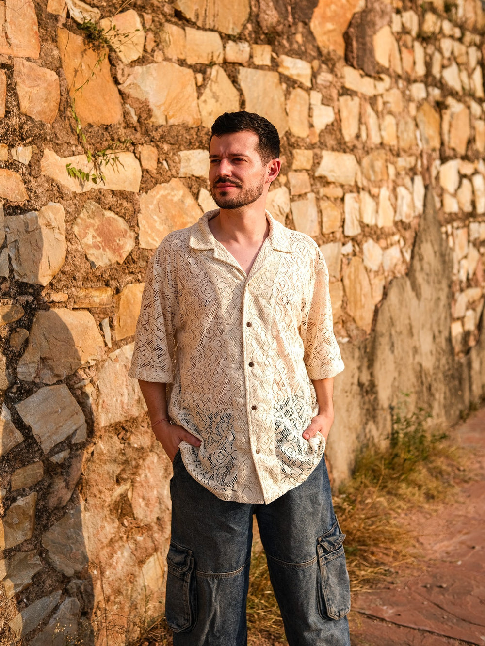 A man dressed in the Anvaya Shirt—crafted from premium mesh net fabric with a refined beige pattern—pairs it with dark cargo pants as he stands hands in pockets, leaning against a sunlit stone wall and gazing off to the side.