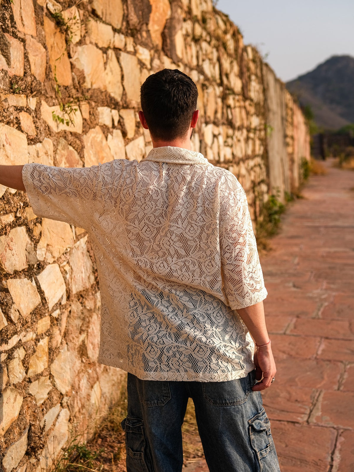 Wearing an Anvaya Shirt crafted from premium mesh net fabric and blue jeans, a person stands with their back to the camera, lightly touching a stone wall along a sunlit path, with mountains rising in the background.