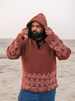 Wearing a Tribal Hoodie in brown, its geometric block print design on full display, someone stands outdoors. They clutch the hood with both hands, while their blue jeans keep them grounded in an open landscape under a pale sky.